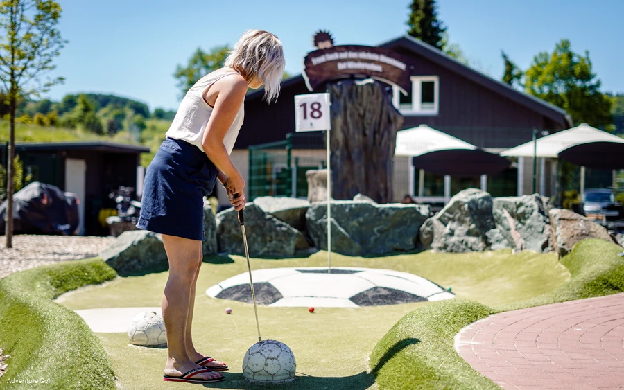 Eine Frau schlägt einen Golfball in Richtung Loch, welches sich auf einem Hügel mit dem Aussehen eines Fußballs befindet.