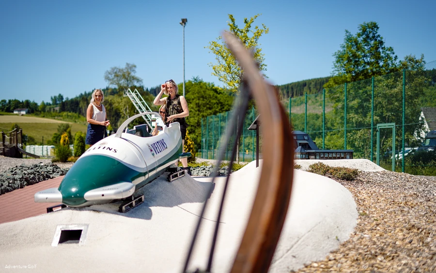Zwei Frauen spielen Golf auf einer Bahn mit einem Bob als Hindernis.